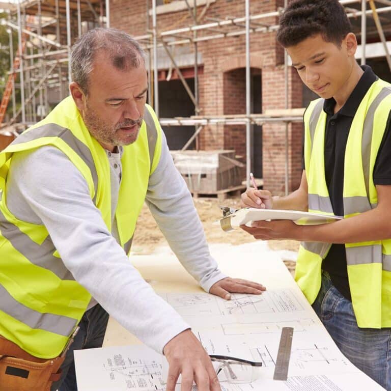 A construction supervisor in a high-visibility vest is reviewing building plans with a younger trainee who is taking notes on a clipboard at an active construction site.